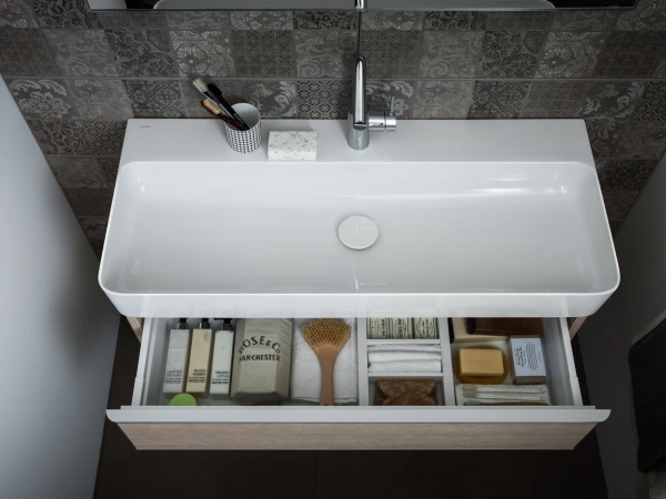 Top-down view of a white bathroom sink with a chrome tap; an open drawer reveals toiletries, towels, a wooden brush and soap against patterned tiles.
