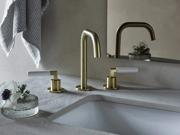 Brass bathroom faucet set with a tall curved centre spout and two lever handles on a speckled grey countertop, with a towel and a glass dish holding a white flower.