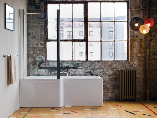 Industrial bathroom with a white freestanding tub and glass screen, exposed brick wall, a large multi-pane window, radiator, and colourful lights.