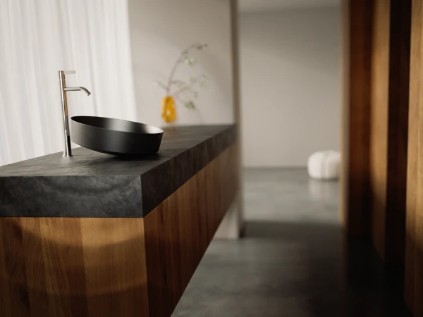 Modern bathroom with a black oval vessel sink on a dark stone worktop, tall chrome tap and wooden cabinetry; blurred background with a yellow vase.
