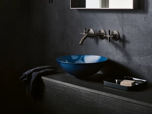 Moody bathroom scene with a blue vessel sink on a black wooden vanity, a wall-mounted silver faucet, dark stone walls, a towel, and a soap tray.