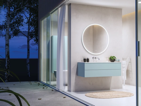 Modern bathroom with a circular backlit mirror above a pale blue floating vanity, concrete walls, and glass doors to a night outdoor space with trees and a woven rug.