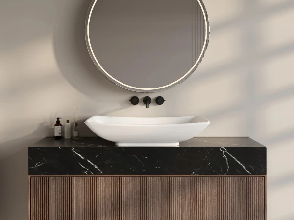 Round backlit mirror above a white vessel sink on a black marble countertop with a wooden slatted cabinet, and bottles to the left.