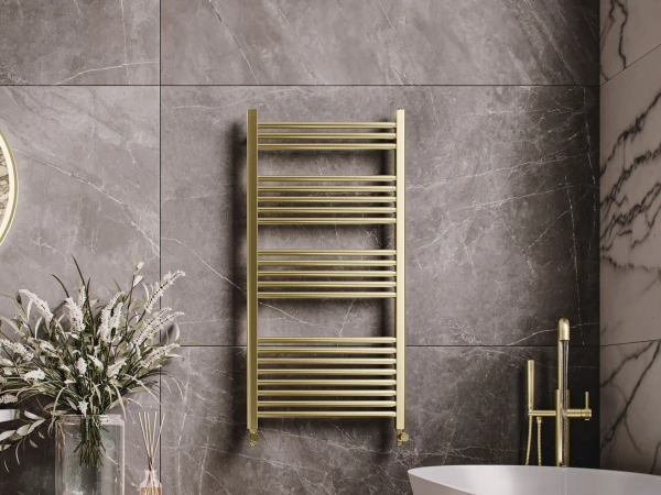 Luxurious bathroom with dark marble walls and floor, a gold towel radiator, a white freestanding tub with brass fixtures, and a wooden slatted cabinet with a vase and diffuser.