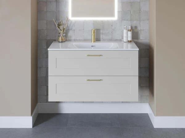 White vanity with two large drawers and gold handles, centre sink beneath a backlit square mirror; grey tiled wall, vase of reeds and toiletries.