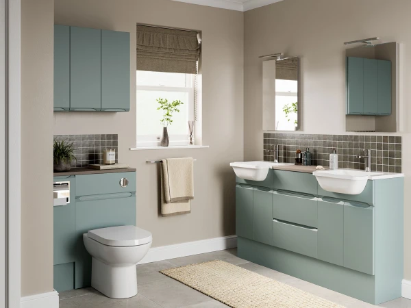 Modern bathroom with blue-green cabinetry, a double-sink vanity, square tiled backsplash, large mirrors, a window with roman shade, plant, towel rail and beige rug.