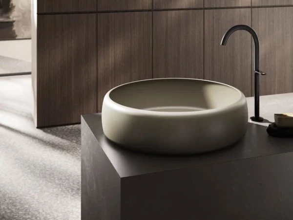 Modern bathroom with a round beige vessel sink on a dark countertop, tall curved black faucet, and wood-paneled wall in soft light.