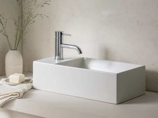 Modern white rectangular bathroom sink with a chrome tap on a pale stone counter, a vase of dried stems, soap and a towel nearby.