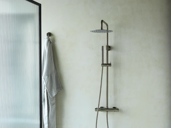 Minimalist bathroom featuring a tall chrome rainfall shower with handheld hose, frosted glass panel, a grey robe hanging, and a white ceramic stool with a bowl of stones.