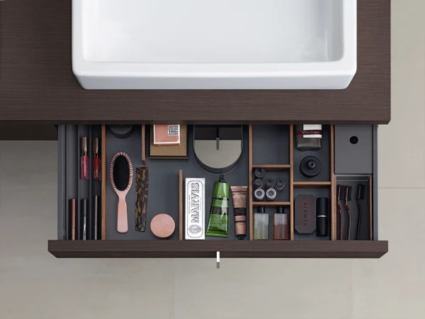 Top-down view of an open dark wood bathroom drawer with compartments filled with cosmetics: brushes, lipstick, nail polish, compacts and bottles.
