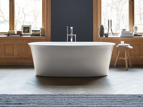 Bright bathroom with a white freestanding tub between large wooden-framed windows and a dark navy divider wall; books, a stool and a blue rug.