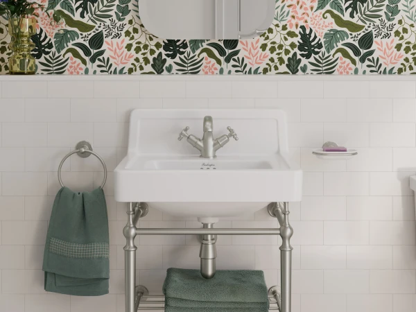 Freestanding white sink on a silver metal frame with a lower shelf and green towels; towel ring, vase of flowers, bold botanical wallpaper, mirrored above, pink hex floor.