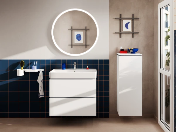 Contemporary bathroom with a round backlit mirror, white two-drawer vanity, blue-tiled wall, towel and toothbrush holder, and a tall white cabinet beside a window.