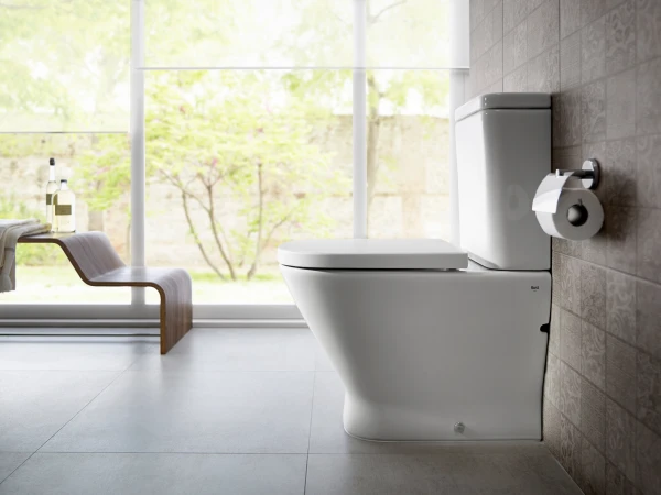 Bright modern bathroom with a white toilet against tiled wall, toilet paper holder, and a wooden bench with towels and bottles by a large window overlooking greenery.