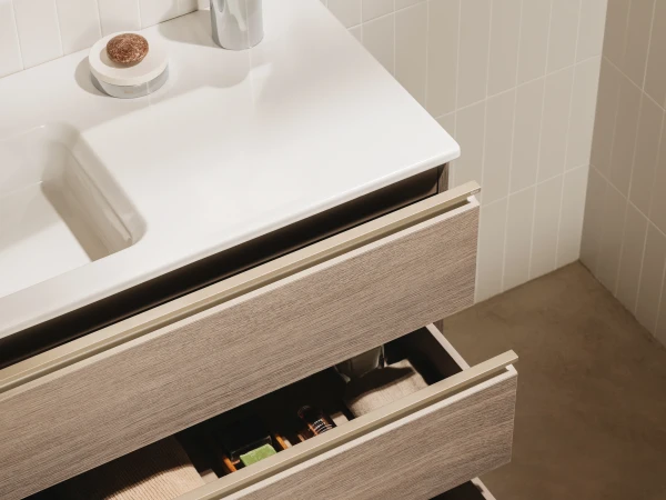 Modern bathroom vanity with pale wood drawers and a white countertop; top holds a toothbrush cup and soap, three drawers open showing towels and toiletries.