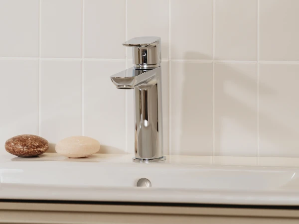 Modern chrome bathroom faucet on a white tiled wall, with a curved mirror edge above and two smooth stones resting on the sink counter.