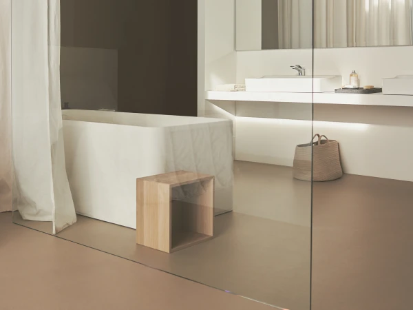 Minimalist bathroom with a white bathtub, sheer curtain, glass divider and large mirrored wall; wooden cube stool and woven basket on a beige floor.