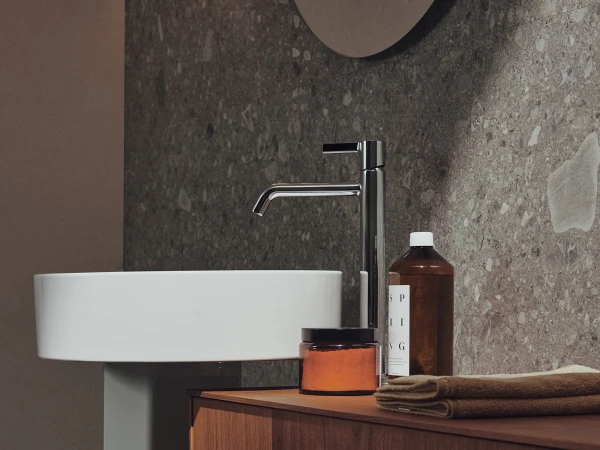 A modern bathroom with a white pedestal sink and chrome tap against a grey speckled stone wall; a wooden vanity holds a brown bottle, an orange jar and a folded towel.