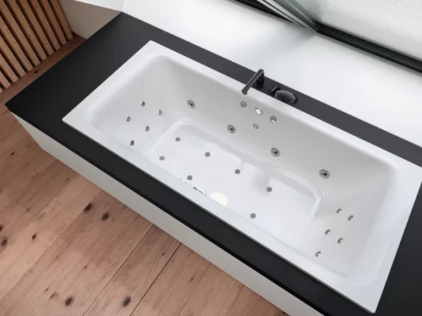 White rectangular Jacuzzi tub with numerous jets, inset into a dark surround on a wooden floor, beside a large window and a wooden slatted wall.