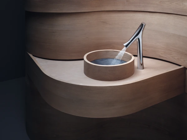 Modern curved wooden bathroom with a circular wooden basin and a sleek chrome tap pouring water into the basin.