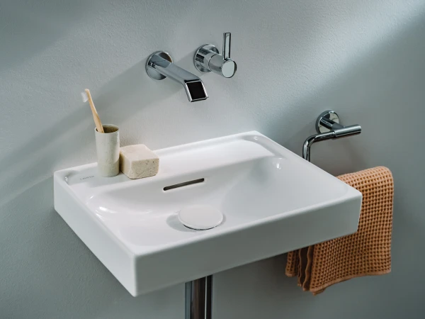 White rectangular sink with chrome wall-mounted tap and round control, beige towel on a chrome rail, toothbrush in a cup and soap block on the sink.