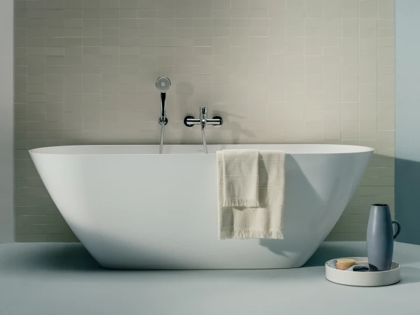 Modern white freestanding bathtub against a beige tiled wall, with a towel over the rim and a blue pitcher on a round tray beside it.