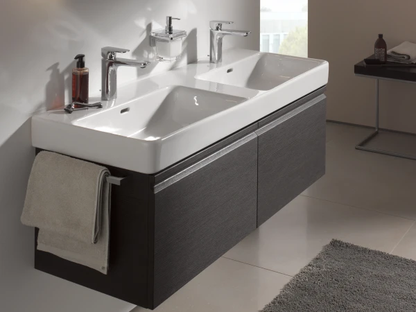 Modern bathroom with a white double sink vanity, chrome taps, and a backlit mirror; dark wood-effect cabinet, beige walls, a grey rug and towel.