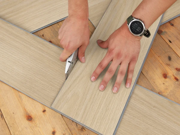 Top-down view of hands cutting a light wood laminate plank with a utility knife during flooring installation; a wristwatch is on one wrist.