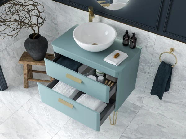 Teal bathroom vanity with round white basin, gold faucet; open drawers reveal towels and toiletries, marble walls, and a wooden stool with a vase.
