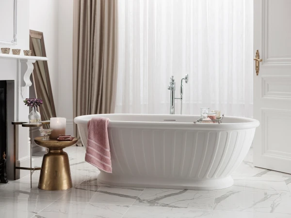 Bright white bathroom with a freestanding ribbed bathtub, pink towel on the rim, marble floor and a gold side table with a candle and flowers; soft curtains in the background.