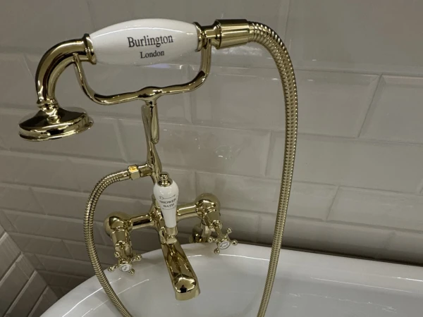 Gold-plated Victorian-style bath faucet with a flexible handheld shower on a white tub, Burlington London label on the shower head, white tiled wall behind.