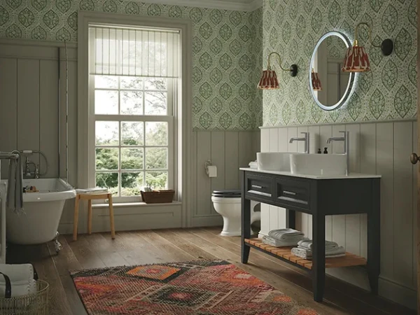 Vintage-style bathroom with green damask wallpaper and wainscoting, freestanding tub by a large window, dark sink vanity, round backlit mirror and red lamps.