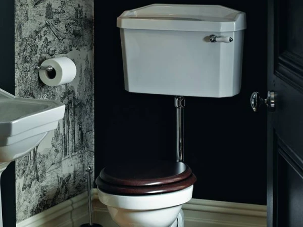 White toilet with a dark wooden seat and chrome fittings in a bathroom, patterned wallpaper on the left and a partial pedestal sink visible.