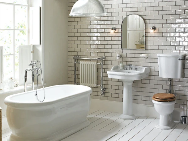 Bright vintage bathroom with a white tub, pedestal sink, mirror, and toilet with a wooden seat; white subway tiles, radiator, and window.