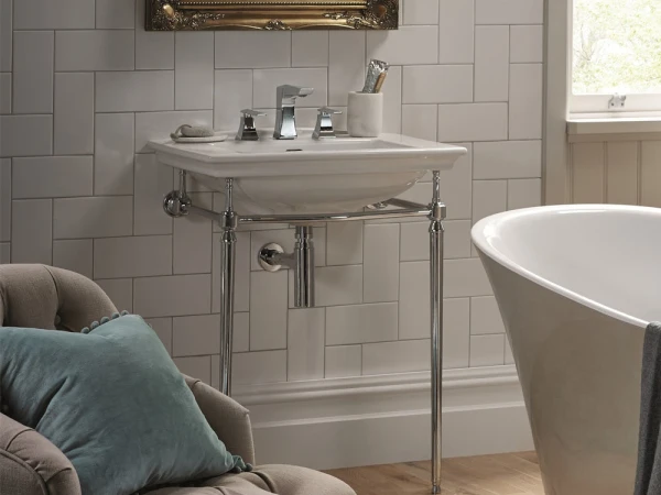 A stylish bathroom with a white sink on chrome legs, a freestanding tub, a tufted beige chair with teal cushions, tiled walls and a window.