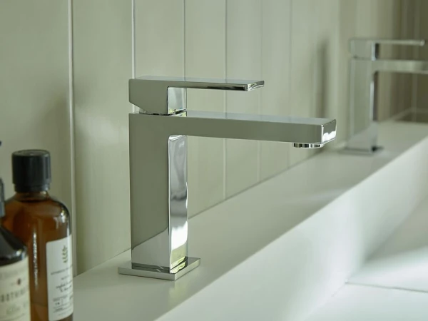Close-up of a sleek chrome rectangular tap on a white sink, with pale tiled wall and brown bottles visible on the left.