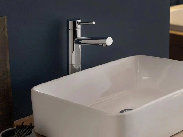 A sleek chrome faucet above a white rectangular sink on a light wood counter, against a dark blue wall; a small dish with cotton swabs sits nearby.