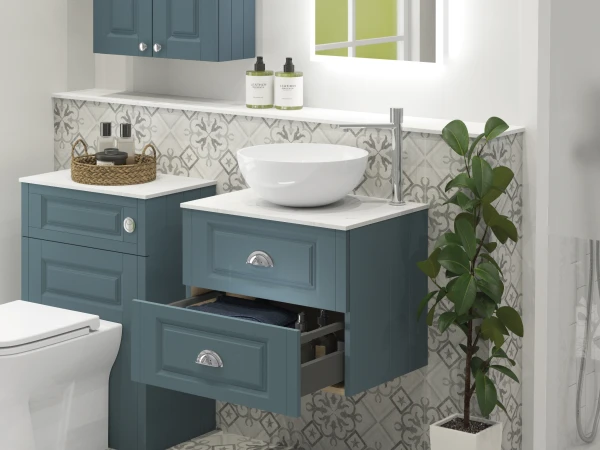 Blue bathroom vanities with white marble tops, round sink, chrome faucet, grey patterned tiles, wicker tray with bottles, drawers, and a potted plant.