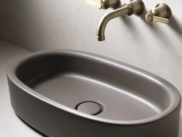 Grey oval basin on a light countertop with a brass wall-mounted faucet and two lever handles, mirror edge visible above on a pale stone wall.