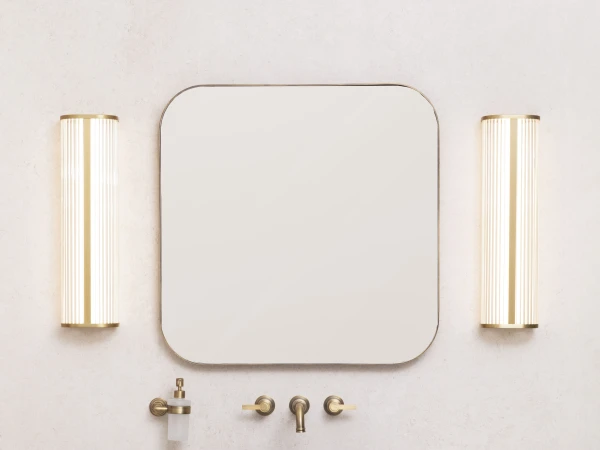 Modern bathroom vanity with a rounded wood cabinet, white countertop, grey vessel sink, brass fixtures, an oval mirror, and vertical wall lights.