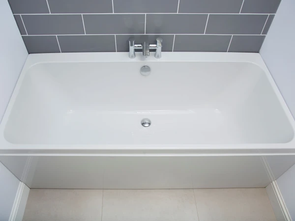 White rectangular bath with chrome taps, set against a grey subway-tiled wall and pale floor tiles.