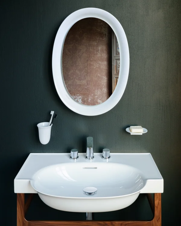 White sink with chrome taps on a wooden stand, an oval white-framed mirror above, toothbrush holder and soap dish on a dark textured wall.