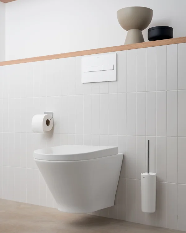 Minimalist bathroom with a wall-hung toilet, white tiled wall, flush plate, toilet paper holder, and a tall brush; decorative bowls on a wooden shelf.