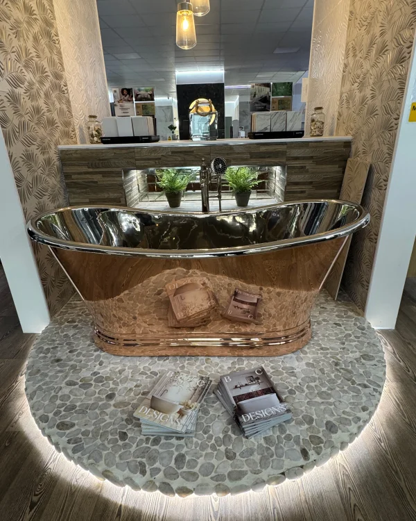 Polished copper freestanding bathtub on a pebble round rug, with magazines and a reception desk with plants in a stylish showroom.