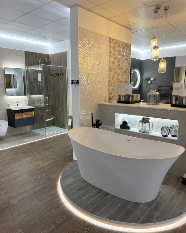 Modern bathroom showroom with a white freestanding tub on a circular lit plinth, grey floor, wall, and a glass shower enclosure labelled Merlyn.