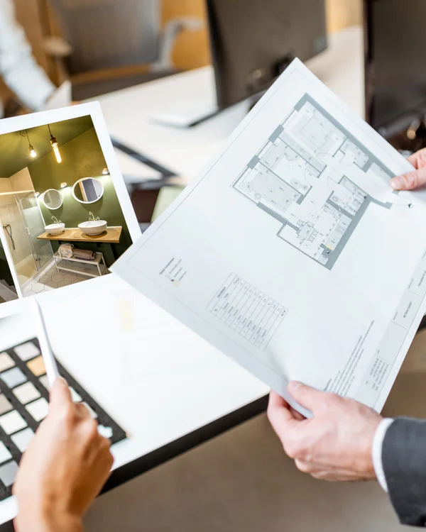 Two people review interior design plans: a photo board showing a green bathroom with round mirrors and two sinks, beside a printed floor plan.