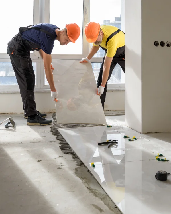 Two construction workers wearing orange hard hats and gloves install large white glossy floor tiles in a partially finished room by a window; spacers and tools on the floor.