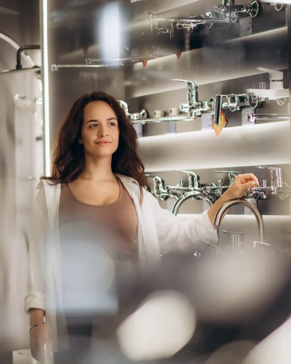 Person with long brown hair stands in a showroom, wearing a taupe top and white shirt, inspecting chrome bathroom taps on display.