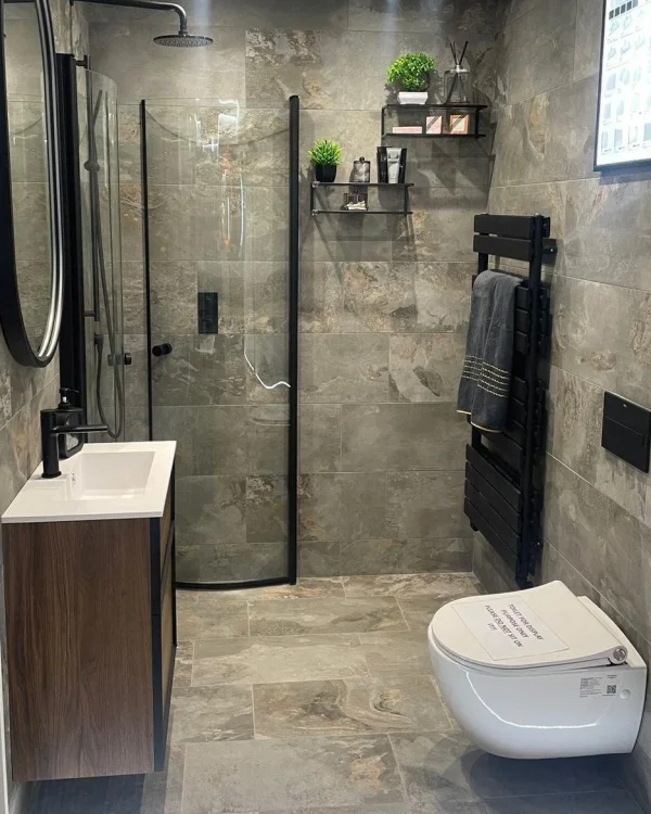 Modern grey-tiled bathroom with a curved glass shower, black-framed mirror, wooden vanity, plant shelves, and a wall-hung toilet with a black towel rail.
