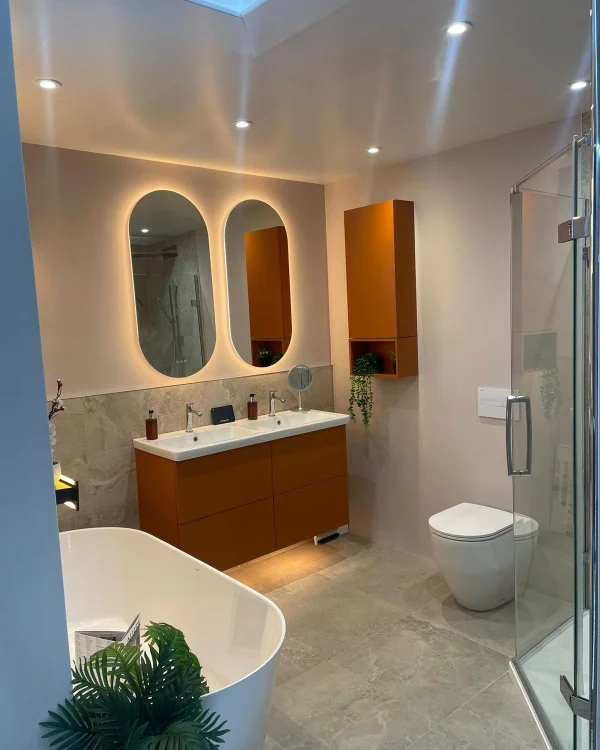 Contemporary bathroom with burnt-orange vanity and wall cabinet, double sinks under backlit oval mirrors, freestanding white tub, glass shower and toilet.